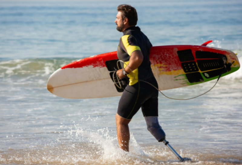 Ein entschlossener Surfer mit einer Prothese am Bein trägt sein Surfboard und läuft ins Meer. Dieses Bild verdeutlicht die Stärke und Möglichkeiten im adaptiven Sport.