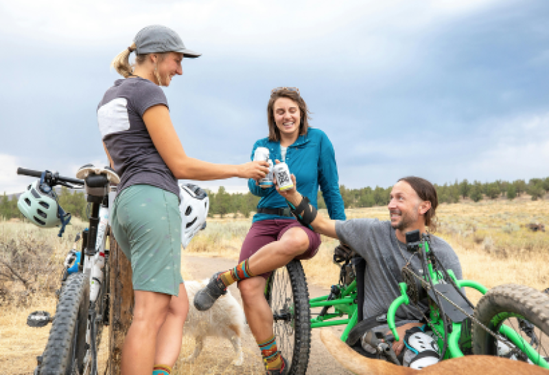 Das Bild zeigt drei Personen, von denen eine eine Beinprothese trägt, während sie während einer Radtour eine Pause einlegen und Wasser teilen. Es vermittelt Inklusion und Freundschaft im Sport.